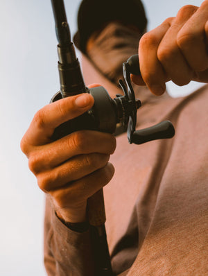 Close-up of a person's hands winding a VoltiQ fishing reel on a rod, with the person wearing a brownish-pink shirt and a dark cap against a pale sky background.