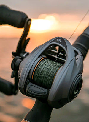 Close-up of a VoltiQ SX baitcasting fishing reel mounted on a handle, with green fishing line wound around the spool and a blurred sunset background.