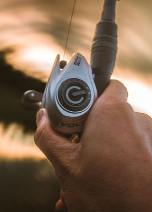 A close-up shot of a person's hand holding a fishing rod with a VoltiQ baitcasting reel in silver and grey. The background is a warm, out-of-focus sunset scene.