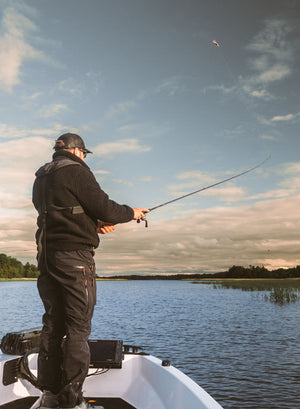 A man stands on the edge of a boat on a lake, casting a fishing rod under a cloudy sky during golden hour.