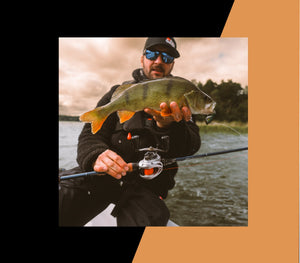 Tobias Fränstam wearing a black cap, sunglasses, and a black fishing jacket holds a freshly caught perch fish while sitting on a boat on the water, with a black and orange graphic border framing the photo.