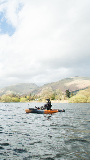A lone angler in a kayak on a relatively calm water, to a backdrop of trees and bushes ascending to mountain tops and a cloudy sky with patches of blue breaking through.
