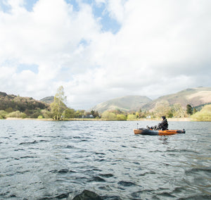 A lone angler in a kayak on a relatively calm water, to a backdrop of trees and bushes ascending to mountain tops and a cloudy sky with patches of blue breaking through.