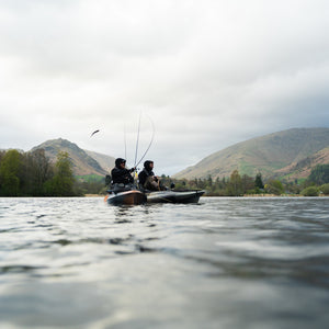 Two people fishing from kayaks on a lake with mountains in the background