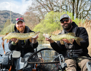 Two anglers holding pike caught on a fishing trip with a scenic background.