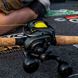 A close-up of an Abu Garcia Low Profile Beast baitcasting fishing reel in black mounted on a cork-finished rod, with bright yellow line spool and part of a person's leg visible in the background on a branded fishing mat.