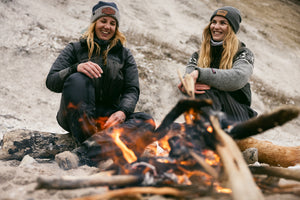 Two ladies sitting by a campfire in a lightly coloured sandy cove landscape