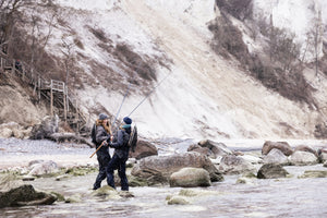 Two anglers stood in the sea, between the rocks, near the beach. In a light landscape with rocks, trees and a wooden staircase up the beach cliff.