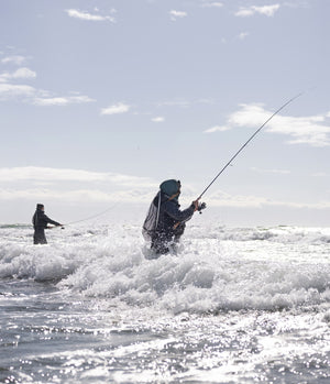 Two people stood fishing in the ocean, with the waves breaking over their thighs, with a clear sky above.