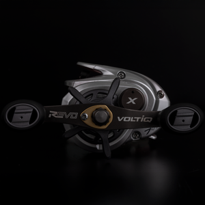 An Abu Garcia VoltiQ X fishing reel with a metallic silver body, black spool arms, and gold accents, featuring the 'REVO' and 'VOLTIQ' branding on the handle, set against a solid black background.