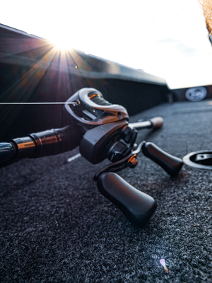 A close-up action shot of a black and silver  Abu Garcia VoltiQ baitcasting reel mounted on a rod, resting on a dark textured carpet inside a boat. The sun is low in the sky, creating a bright flare and backlit effect.