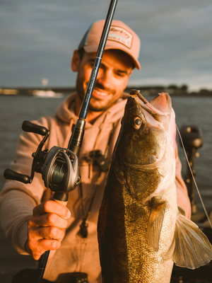A man in a beige hoodie and cap holds a large freshly caught fish while holding a fishing rod with an Abu Garcia VoltiQ baitcasting reel on a boat, with water and a cloudy sky in the background.