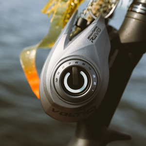 Close-up of a silver and black VoltiQ fishing reel with a power button dial, attached to a rod, with a soft orange and yellow lure visible behind it against a blurred water background. 