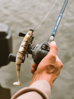 A close-up of a person's hand holding a fishing rod with an  Abu Garcia VoltiQ SX baitcasting reel, with a soft plastic fish-shaped lure dangling from the line. The lure has a realistic fish pattern in beige, brown, and black, and the rod is dark blue with white branding.