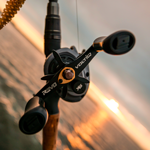 Close-up of a black and gunmetal Abu Garcia VoltiQ SX baitcasting reel mounted on a rod, with a blurred sunset over water in the background.