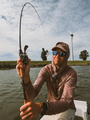 A man wearing a brown hooded long-sleeve shirt, sunglasses, and a cap casting a fishing rod with an  Abu Garcia VoltiQ fishing reel while sitting on a boat on the water, with a wind turbine and trees in the background.