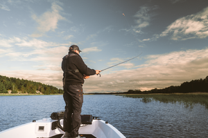A man in black fishing apparel stands on the bow of a boat, casting a fishing rod out over a calm lake under a cloudy sky.